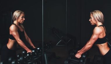 woman staring in mirror at gym holding weights, gym addiction