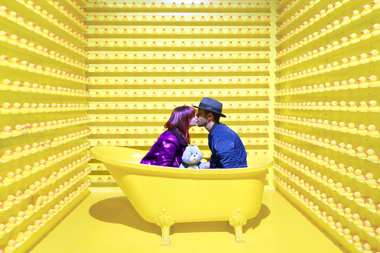 A couple holding a Care Bear kisses in a bathtub surrounded by rubber ducks