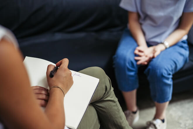 A therapist takes notes as she listens to another woman on a couch