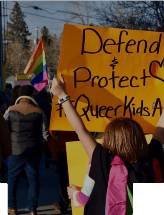 LGBTQ rally with a sign that says "Defend & Protect Queer Kids"